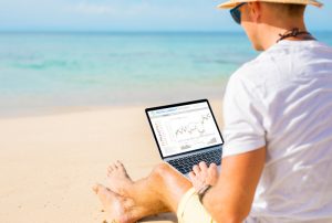 Ein Mann sitzt mit Laptop am Strand und handelt mit Aktien.